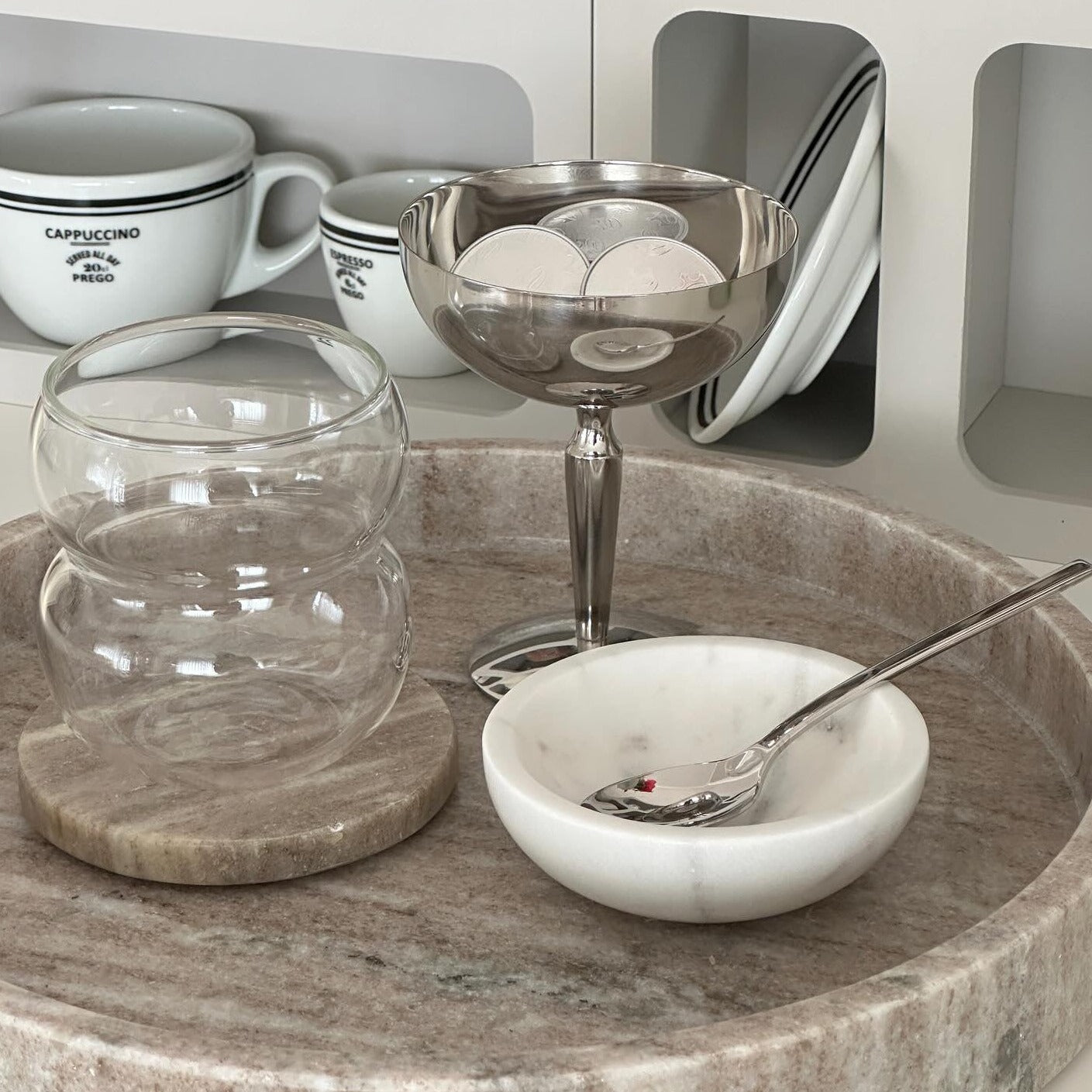 Ein stilvolles, modernes Bubble Glas | Parisian Love, präsentiert auf einer Servierplatte mit einem Untersetzer. Daneben stehen ein Cappuccino- und ein Espressoglas. Luxuriöses Trinkglas mit zeitlosem, gewölbtem und welligem Design sowie ästhetischem Appeal. Handgefertigt und mundgeblasen für ein einzigartiges Trinkerlebnis. Perfekt für Cocktails, Kaffee oder Erfrischungsgetränke. Ein Blickfang auf jedem Tisch. Ideal für den täglichen Gebrauch oder besondere Anlässe. Erleben Sie Luxus und Eleganz.