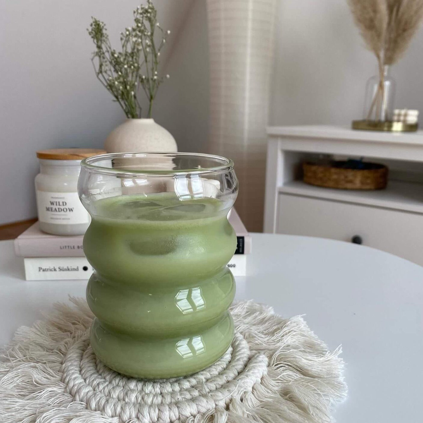 Ein stilvolles Bubble Glas | Venice Dream, serviert auf einem Tisch mit einem erfrischenden Matcha Latte und Eiswürfeln. Im Hintergrund befinden sich zwei Bücher und eine Kerze. Luxuriöses Trinkglas mit einem einzigartigen, gewölbten Bubble Design und ästhetischem Appeal. Handgefertigt für ein einzigartiges Trinkerlebnis mit heißen oder kalten Getränken. Ideal für den täglichen Genuss oder besondere Anlässe. Erleben Sie Eleganz und Gemütlichkeit mit diesem hochwertigen, einzigartigen Glas.