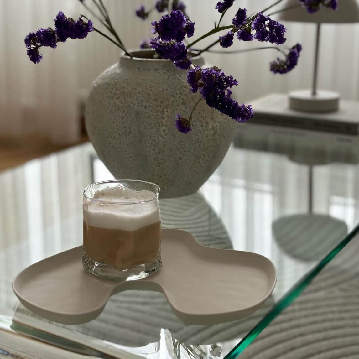 Ein ästhetisches Diamond Twist Glas, präsentiert auf einem Tisch mit einem köstlichen Latte Macchiato darin. Das Glas steht auf einer Servierplatte, dahinter befindet sich eine Pflanze. Luxuriöses Trinkglas mit markantem Design und ästhetischem Appeal. Handgefertigt für ein exquisites Trinkerlebnis. Perfekt für Cocktails, Whiskey oder erfrischende Getränke. Erleben Sie Luxus und Eleganz mit diesem hochwertigen, einzigartigen Glas. Stilvoll und zeitlos gestaltet für anspruchsvolle Genießer.