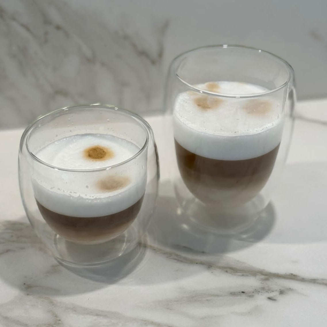 Unser Double Wall Glas 350ml mit doppelwandigem Design, gefüllt mit Cappuccino, auf einem Tisch. Dieses hochwertige, stilvolle und modern designte Trinkglas bietet ein einzigartiges, ästhetisches Trinkgefühl. Perfekt für kalte und heiße Getränke, wie Kaffee und Cappuccino. Ein attraktiver und edler Blickfang, der Luxus und Eleganz in jede Umgebung bringt. Ideal für den täglichen Genuss oder besondere Anlässe.