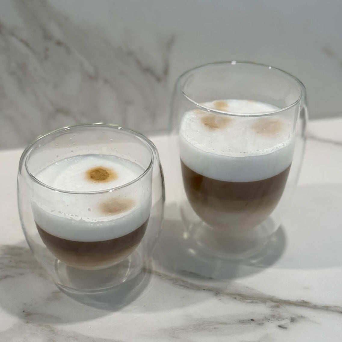 Unser Double Wall Glas 250ml mit doppelwandigem Design, gefüllt mit Cappuccino, auf einem Tisch. Dieses hochwertige, stilvolle und modern designte Trinkglas bietet ein einzigartiges, ästhetisches Trinkgefühl. Perfekt für kalte und heiße Getränke, wie Kaffee und Cappuccino. Ein attraktiver und edler Blickfang, der Luxus und Eleganz in jede Umgebung bringt. Ideal für den täglichen Genuss oder besondere Anlässe.