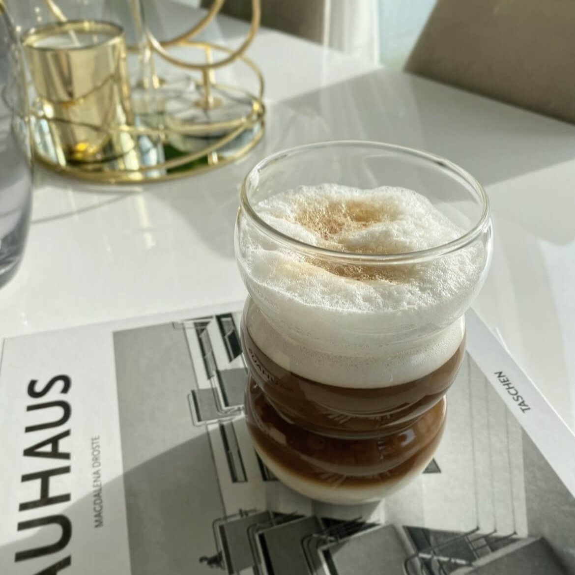 Cappuccino serviert in einem modernen Bubble Glas | Ibiza Sunkissed auf einem Buch mit einem Untersetzer - Luxuriöse, ästhetische Glasware für erfrischende und heiße Getränke. Einzigartiges Bubble-Design und stilvolle Linienstruktur verleihen dem Glas einen modernen und stylischen Look. Handgefertigt und spülmaschinenfest. Ideal für Blogger, Influencer und Feierlichkeiten. Genieße deinen Matcha Latte oder Mojito mit Stil und Luxus.