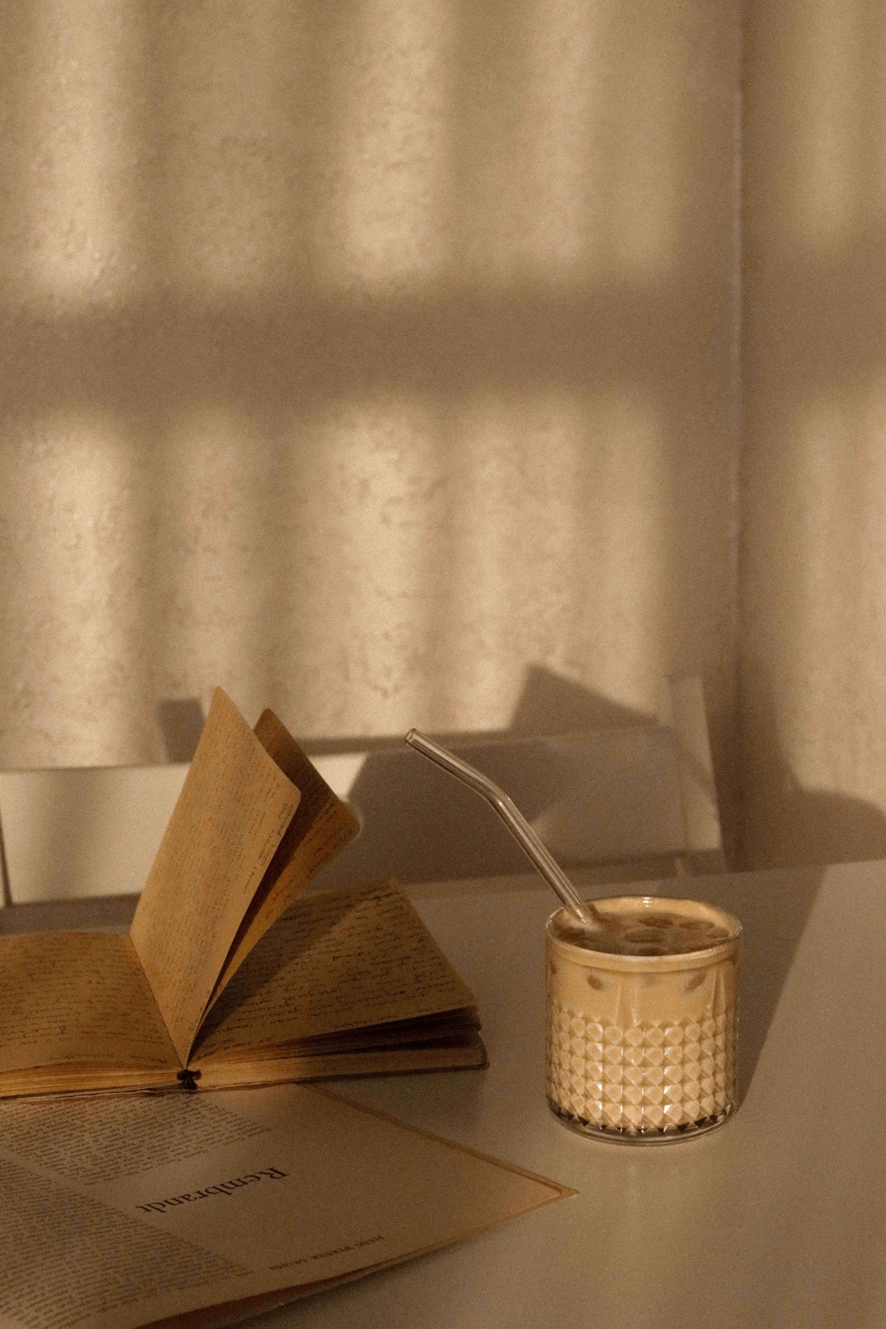 Auf diesem Bild ist ein Glas mit Kaffee zusehen, worin unser Strohhalm aus Glas ist. Daneben liegt ein Buch.